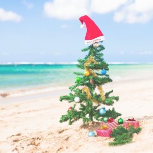 geschmückterWeihnachtsbaum mit Weihnachtsmütze und Geschenken an einem Strand mit türkisblauem Meer im Hintergrund zum Thema Weihnachten im Sommer