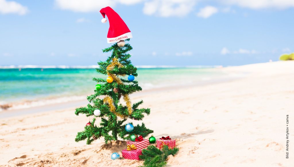 geschmückterWeihnachtsbaum mit Weihnachtsmütze und Geschenken an einem Strand mit türkisblauem Meer im Hintergrund zum Thema Weihnachten im Sommer