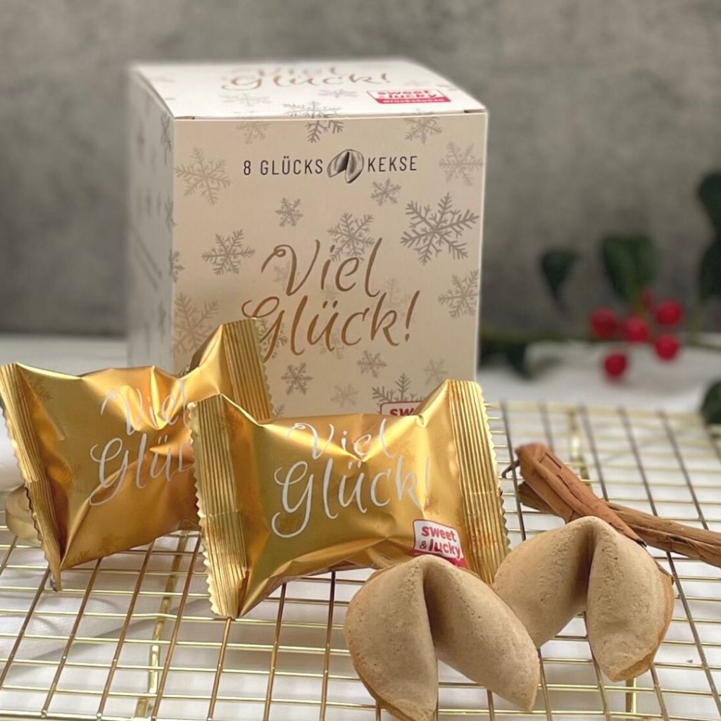Weihnachtsglückskekse: Im Hintergrund die Faltschachtel mit der Aufschrift "Viel Glück", zwei einzeln in Goldfolie eingepackte Glückskekse und zwei ausgepackte Glückskekse.